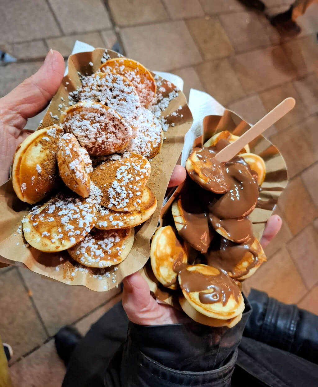 Two cardboard trays filled with small round pancakes.  One has sugar and cinnamon on them, the other has chocolate sauce