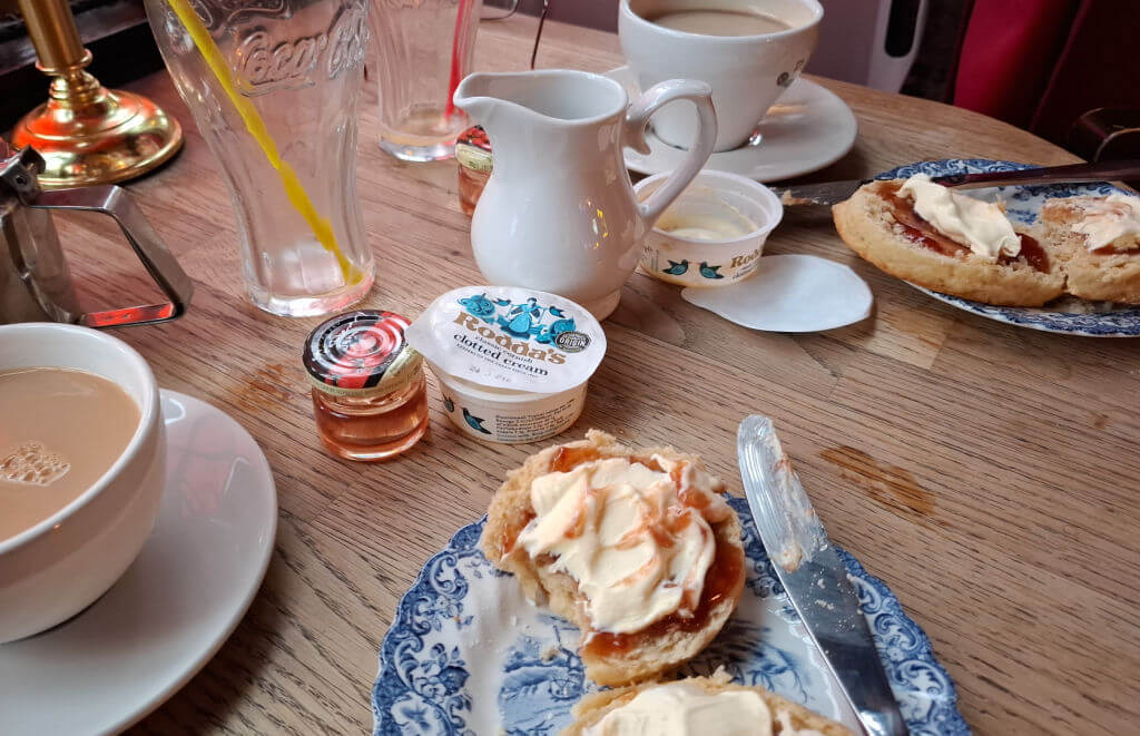 Two plates with scones with cream and jam on a table next to cups of tea and a white jug of milk