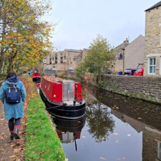 A red-painted canal boat on a canal. A woman in a blue coat with a blue backpack is walking on the towpath, and buildings across the canal are reflected in the water