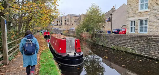 A red-painted canal boat on a canal. A woman in a blue coat with a blue backpack is walking on the towpath, and buildings across the canal are reflected in the water
