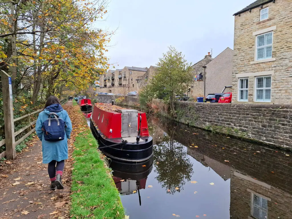 A red-painted canal boat on a canal. A woman in a blue coat with a blue backpack is walking on the towpath, and buildings across the canal are reflected in the water