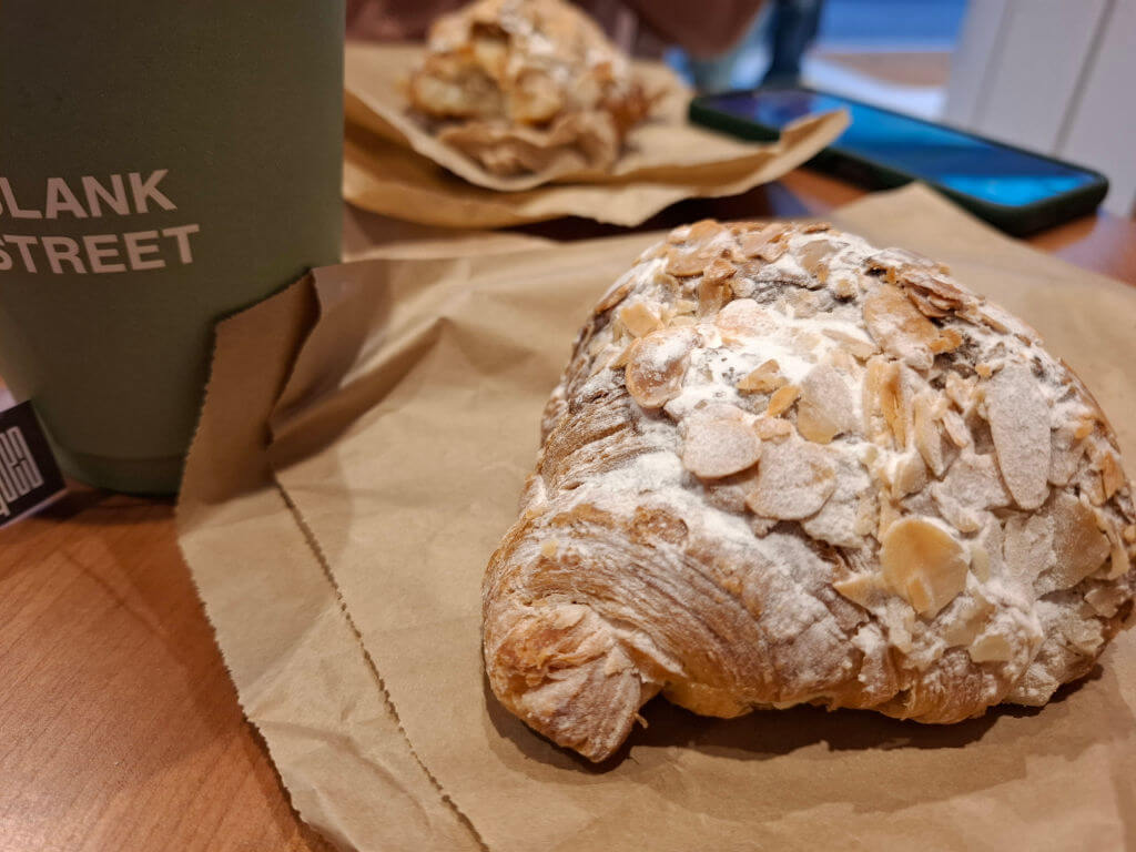 A large almond-encrusted croissant on a wooden cafe table next to a cardboard takeaway cup of tea