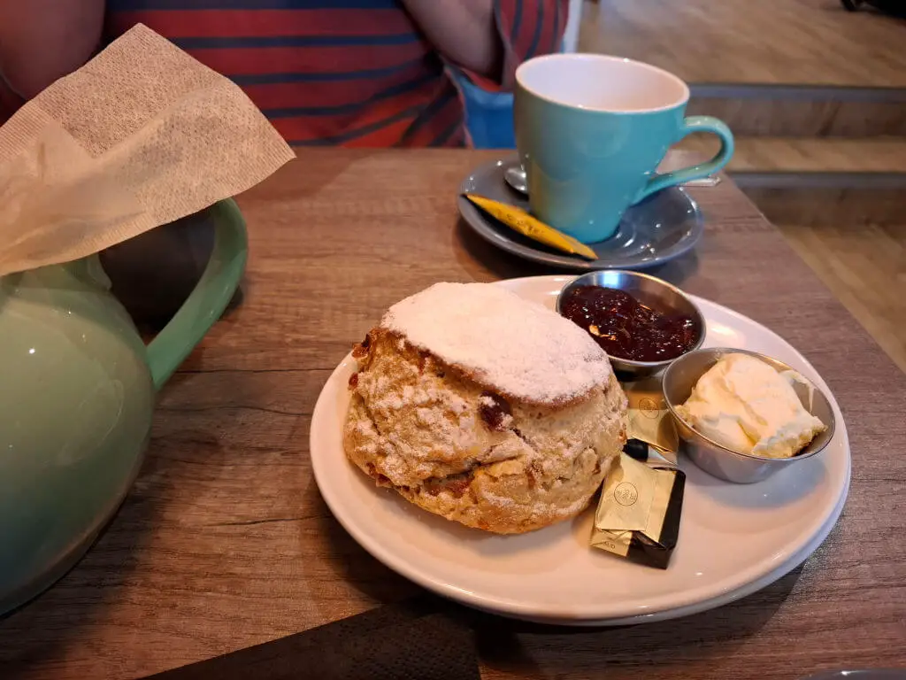 A huge scone with icing sugar on the top sits on a plate with butter, cream and jam on a cafe table. Behind it is a turquoise blue teacup on a saucer