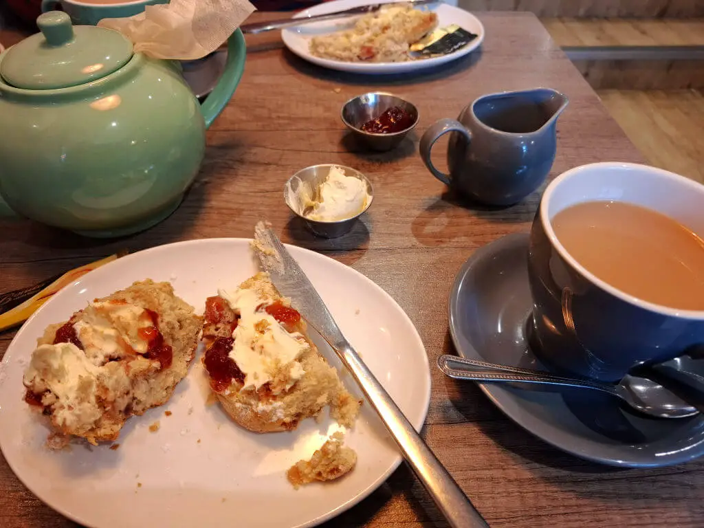 Half of a huge scone with cream and jam on a plate with a knife, and the remains of the jam and cream in small pots on a table next to a cup of tea