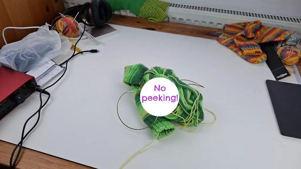 A cream board on a wooden table with a green striped sock, more orange multi-striped socks, balls of yarn and a set of headphones lying on it.