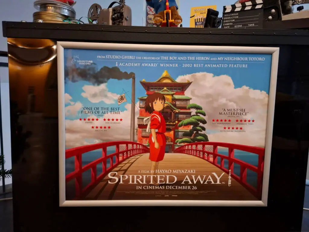 A cinema poster for the film Spirited Away, showing a girl in red clothing standing on a bridge in front of a Japanese bath house