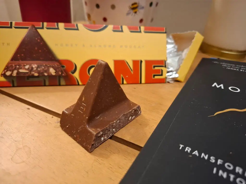 A piece of triangular Toblerone chocolate on a wooden coffee table next to a book with a black cover