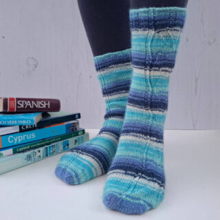 A pair of striped socks in shades of blue and ecru modelled on feet against a white painted board background. Behind the socks is a pile of travel books.