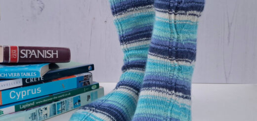 A pair of striped socks in shades of blue and ecru modelled on feet against a white painted board background. Behind the socks is a pile of travel books.