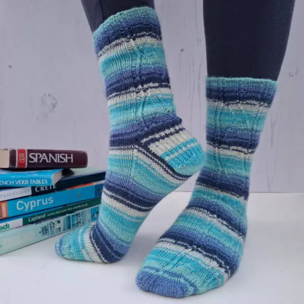 A pair of striped socks in shades of blue and ecru modelled on feet against a white painted board background. The model is standing in such a way as to show off the heel of the sock. Behind the socks is a pile of travel books.