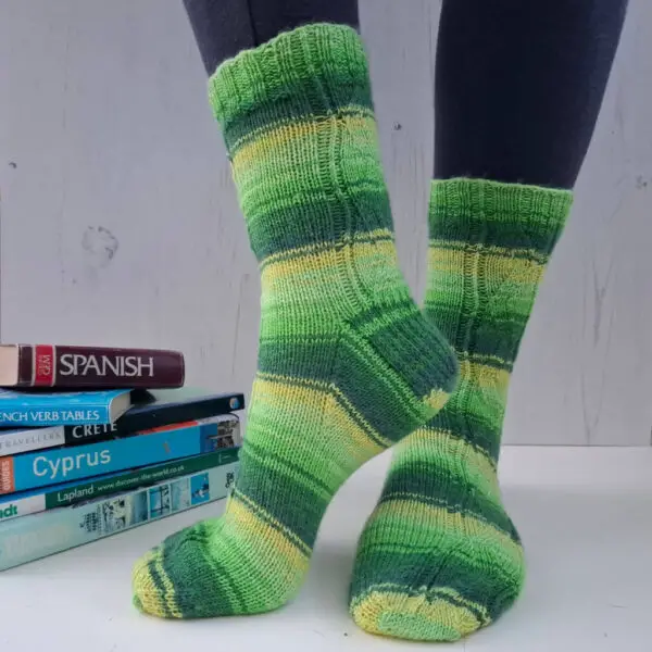 A pair of striped socks in shades of green and yellow modelled on feet against a white painted board background. Behind the socks is a pile of travel books.