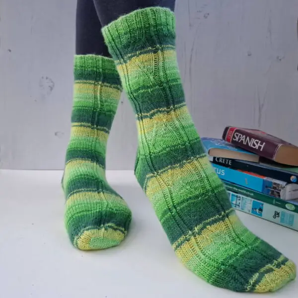 A pair of striped socks in shades of green and yellow modelled on feet against a white painted board background. Behind the socks is a pile of travel books.