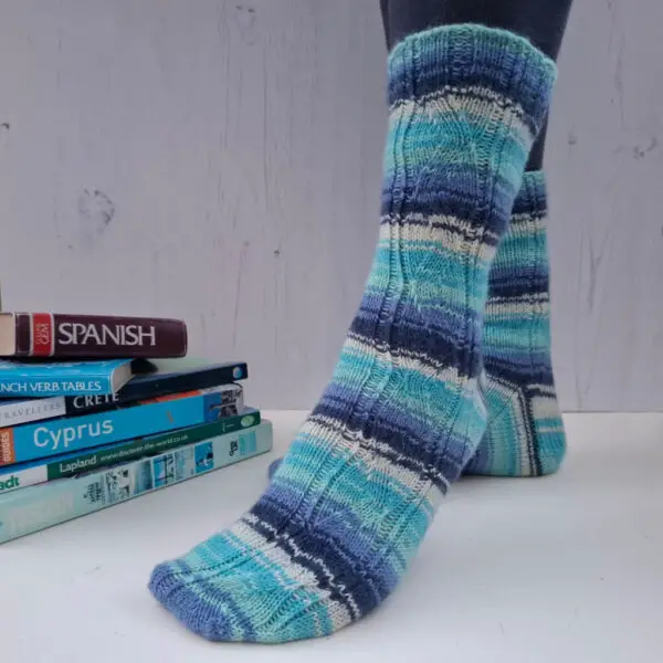 A pair of striped socks in shades of blue and ecru modelled on feet against a white painted board background. Behind the socks is a pile of travel books.