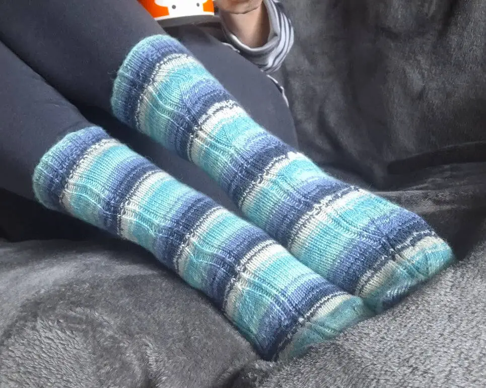 A pair of striped socks in shades of blue and ecru modelled on feet. The model is sitting on a grey fleece blanket on a sofa.