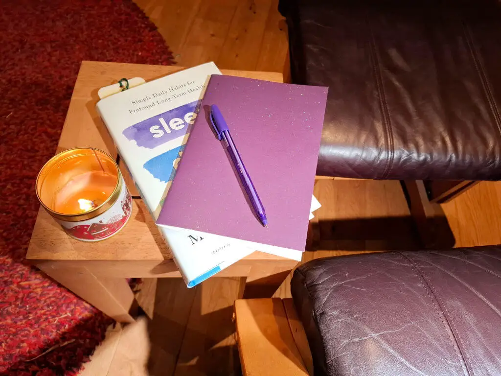 A purple notebook resting on a hardback book on a wooden coffee table next to a lit candle in a tin