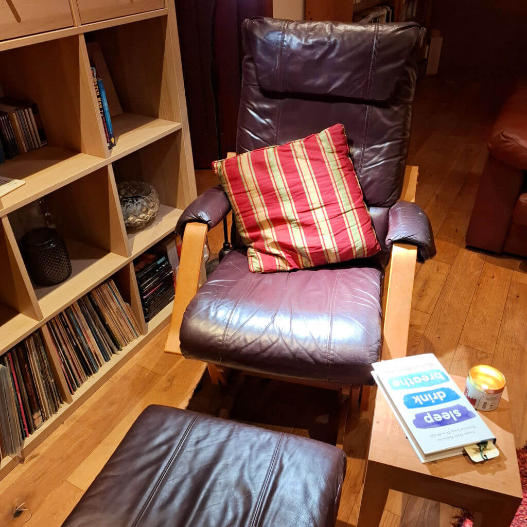 A brown leather rocking chair with matching footstool next to a shelving unit. There is a red and gold striped cushion on the chair