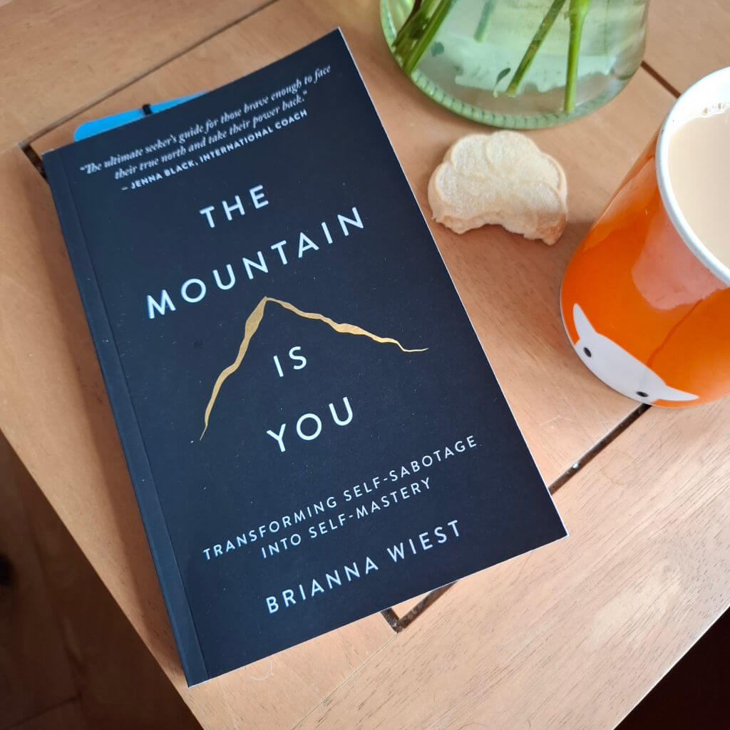 A book with a black cover is lying on a wooden coffee table next to an orange mug of tea, a half-eaten shortbread biscuit and a vase of flowers