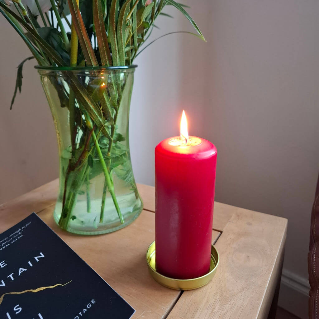 A tall red candle standing on a coffee table next to a book with a black cover and a vase of flowers. The candle is lit.
