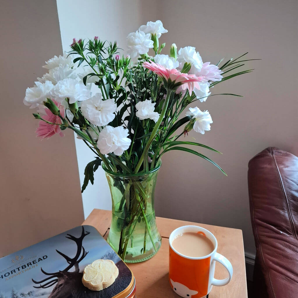 A vase of pink and white flowers in a green glass vase standing on a wooden coffee table next to an orange mug of tea and a metal shortbread tin with a biscuit on the top