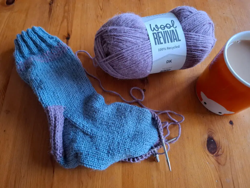 A partly-knitted blue and pink sock lying on a wooden table next to an orange mug of tea