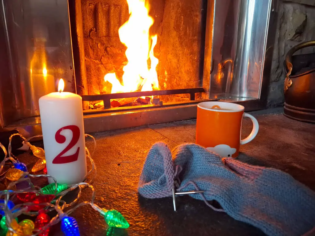 An orange mug of tea on a stone hearth next to a partly-knitted sock, a tall candle with the number 2 on it and fairy lights. There is a fire blazing in the background