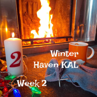 An orange mug of tea on a stone hearth next to a partly-knitted sock, a tall candle with the number 2 on it and fairy lights. There is a fire blazing in the background