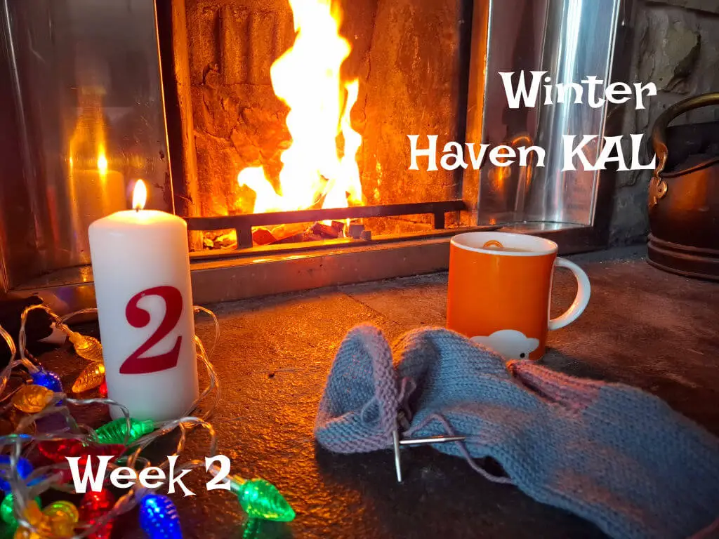 An orange mug of tea on a stone hearth next to a partly-knitted sock, a tall candle with the number 2 on it and fairy lights. There is a fire blazing in the background