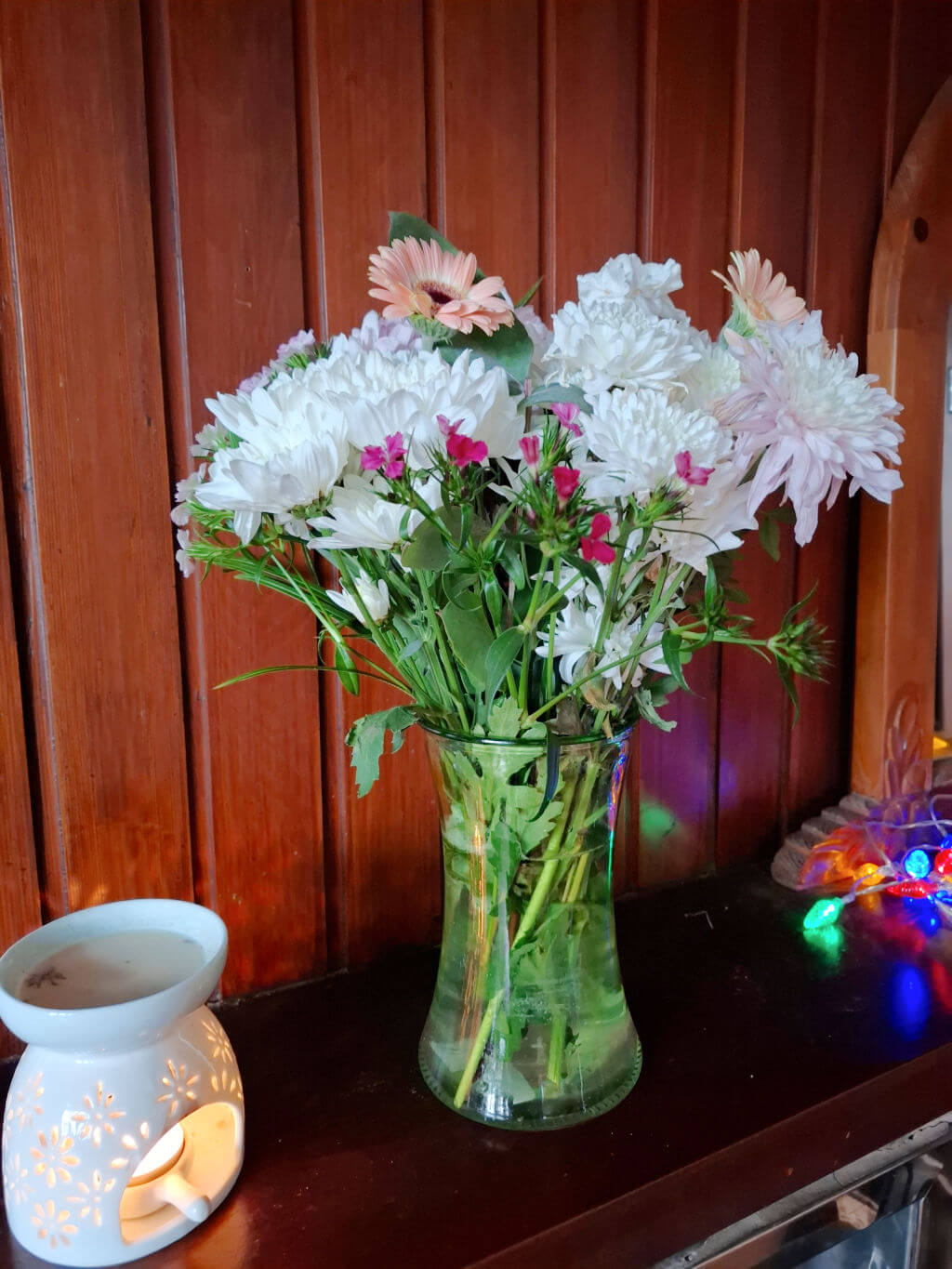A vase of flowers in pink and white on a wooden mantelpiece next to a wax melt burner and a string of coloured fairy lights