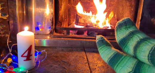 A pair of feet resting on a stone hearth modelling green striped socks, a tall candle with the number 4 on it and fairy lights. There is a fire blazing in the background.