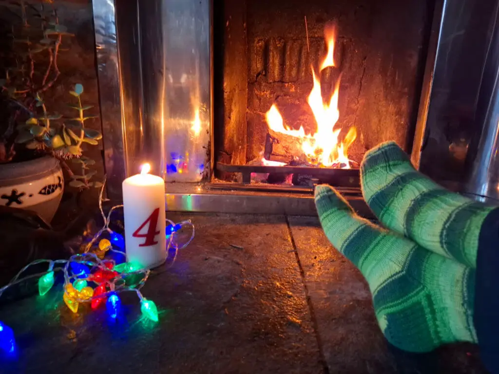 A pair of feet resting on a stone hearth modelling green striped socks, a tall candle with the number 4 on it and fairy lights. There is a fire blazing in the background.