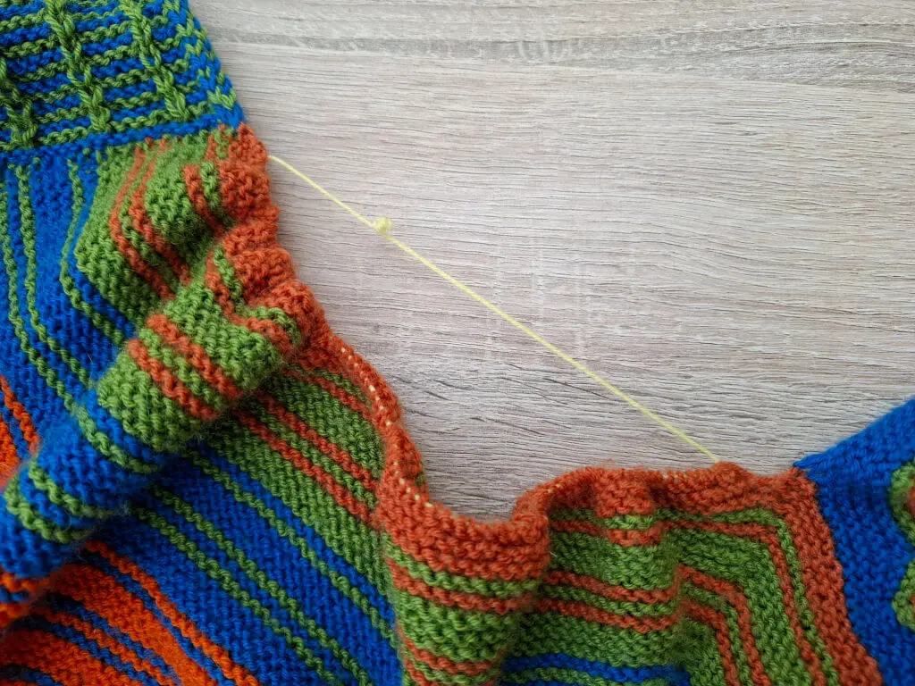 Close-up of colorful knit fabric with blue, green, and orange stripes. A thin yellow thread runs across the wooden table background, creating contrast.