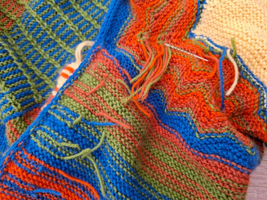 Close-up of colorful knitting with vibrant blue, orange, green, and yellow patterns. A needle is threaded through, suggesting an ongoing project.