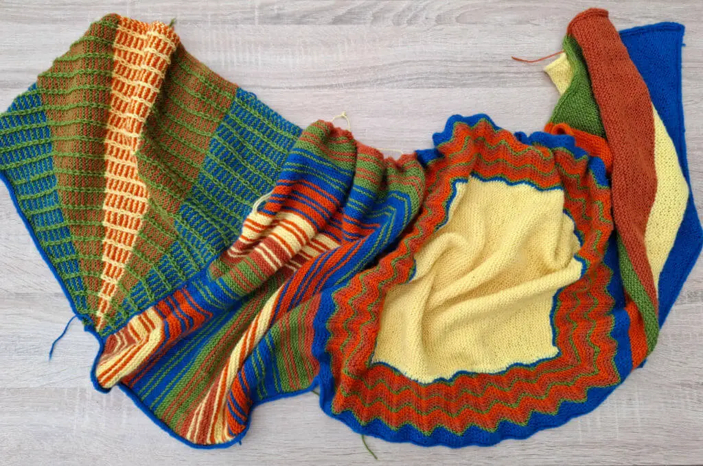 A colorful knitted shawl is spread on a light wooden surface, featuring patterns in green, blue, orange, yellow, and brown stripes with scalloped edges.