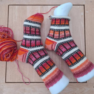 Two colorful knitted socks with geometric patterns in orange, red, and black are displayed on mannequin feet. A ball of matching yarn sits nearby.