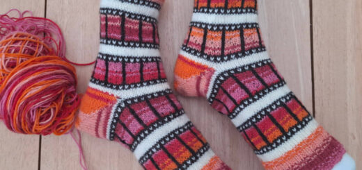 Two colorful knitted socks with geometric patterns in orange, red, and black are displayed on mannequin feet. A ball of matching yarn sits nearby.