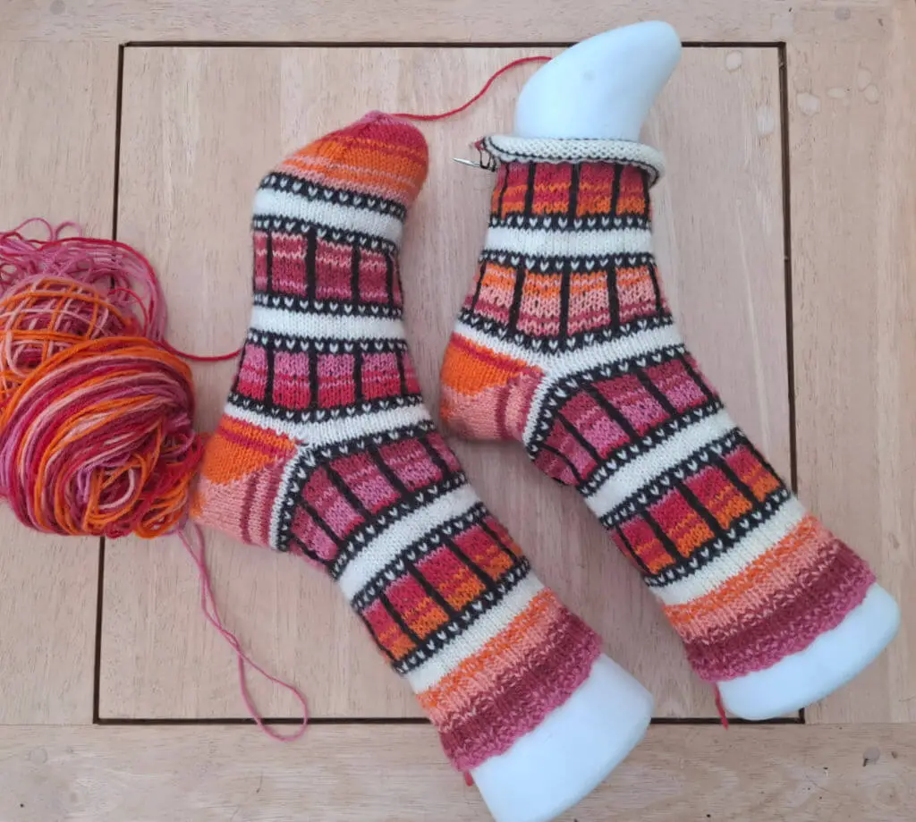 Two colorful knitted socks with geometric patterns in orange, red, and black are displayed on mannequin feet. A ball of matching yarn sits nearby.