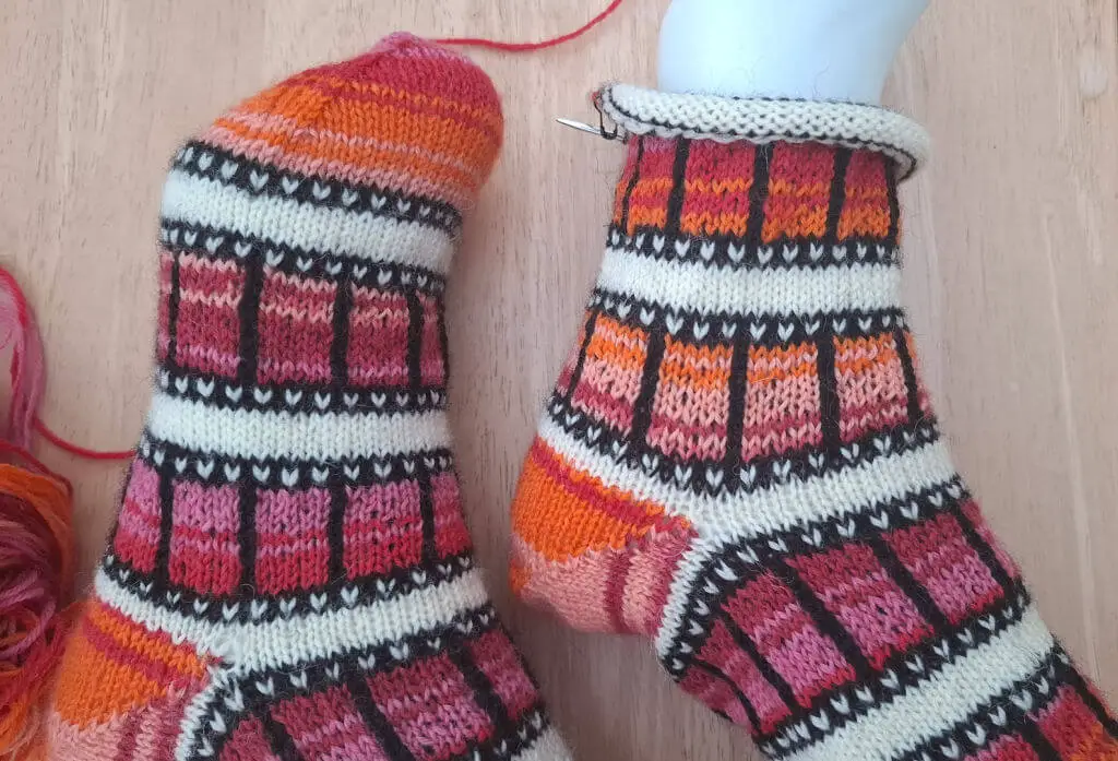 Colorful knitted socks with a bold geometric pattern in shades of orange, pink, and black. One sock in progress on knitting needles. Warm and cozy tone.