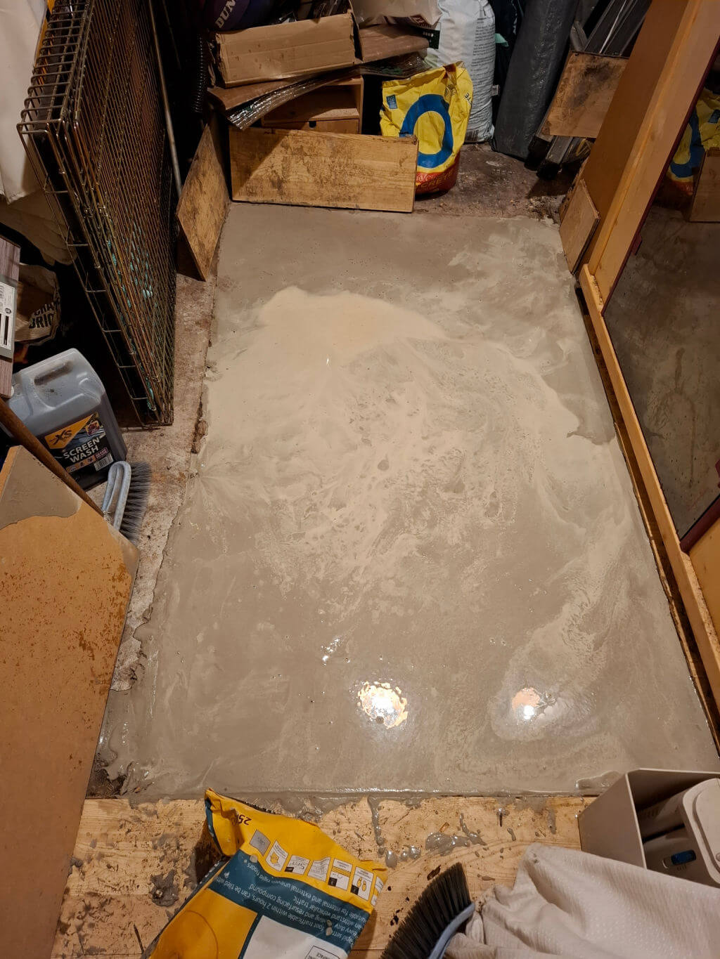 A newly poured concrete floor in a cluttered room. Reflections of lights create a smooth surface effect. Surrounding materials include cardboard, wood, and tools.