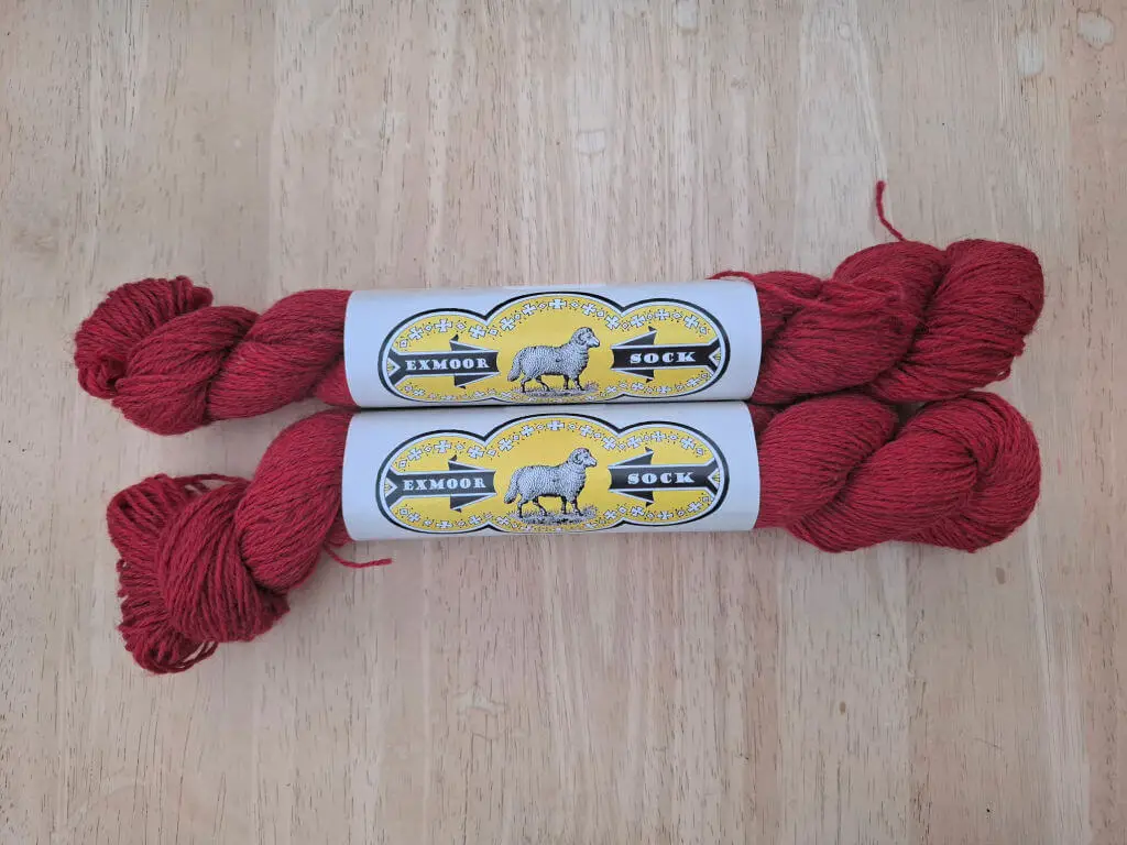 Two skeins of deep red yarn labeled "Exmoor Sock" lie side by side on a wooden surface. The labels feature a sheep illustration on a yellow background.