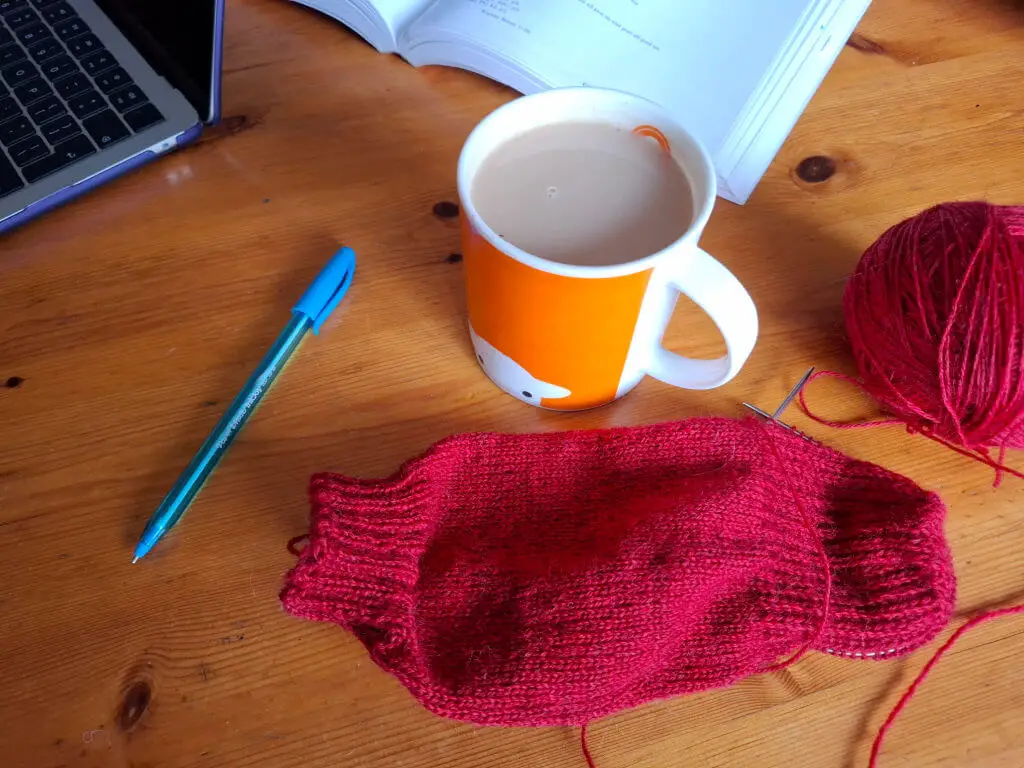 A cozy scene with a laptop, an open book, a bright mug of coffee, a blue pen, and red knitting on a wooden table, conveying a warm, creative vibe.