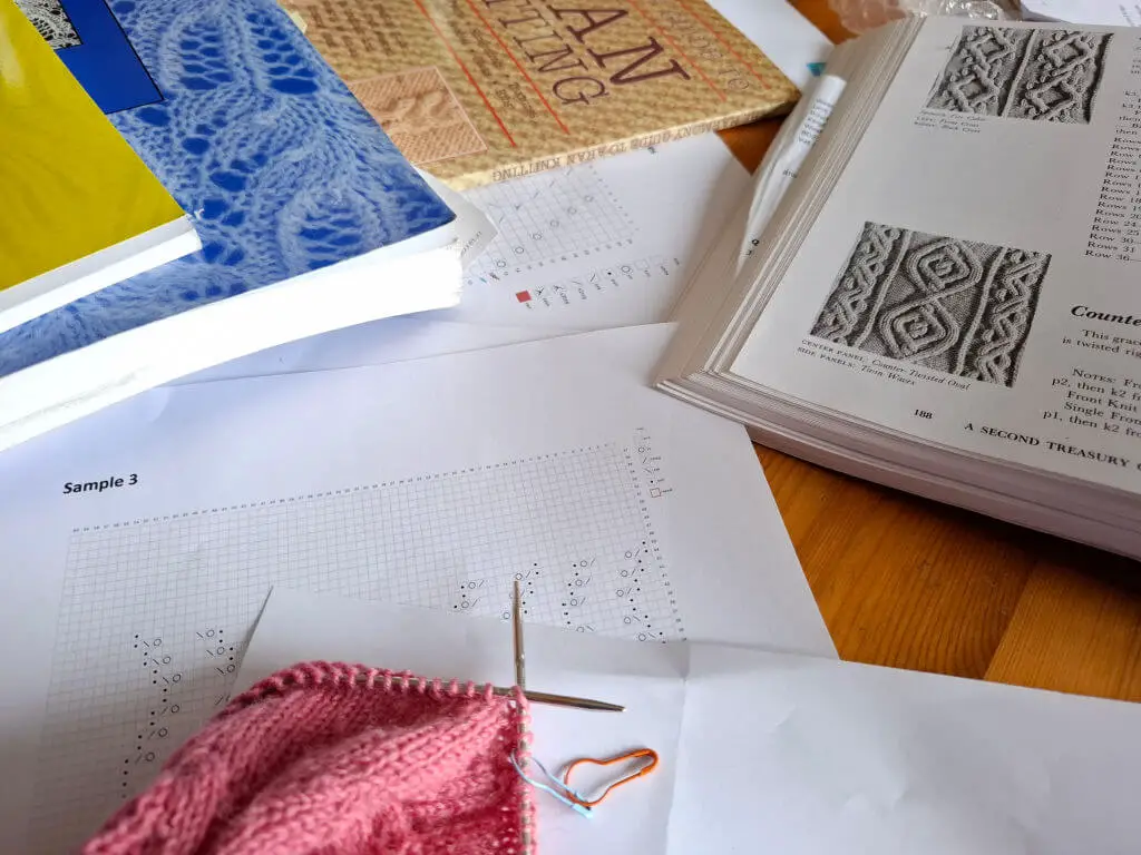 Open knitting books with intricate lace patterns and charts on a wooden table. A pink knitted piece with attached needles and an orange stitch marker lies on top.