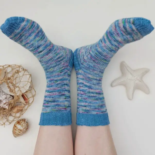 Two feet wearing colourful knit socks rest on a white surface, surrounded by seashells, a fishnet, and a starfish, evoking a beachy vibe. Winwick Mum | Basic 4ply Socks | Helical knitting | Pooling and flashing in yarn