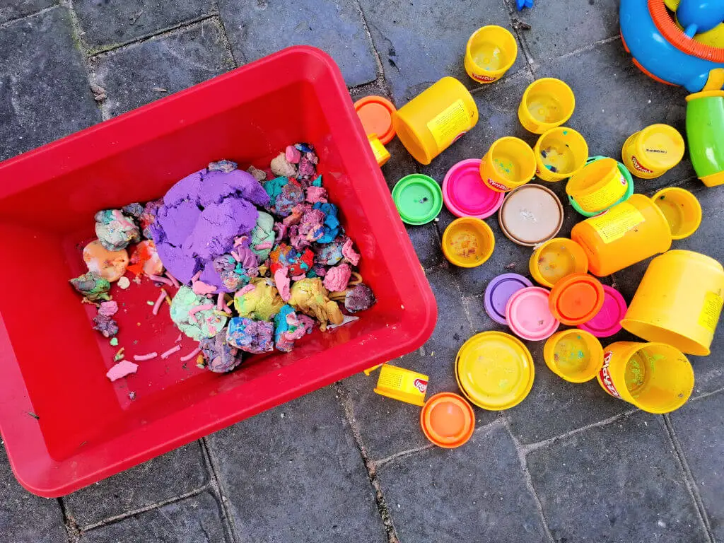 A red bin filled with mixed, colorful Play-Doh on stone pavement. Empty yellow Play-Doh containers and lids are scattered beside it, suggesting playful creativity.