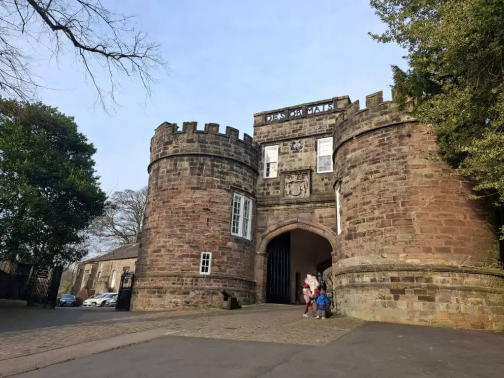 Stone castle entrance with two round towers under a clear sky. People walk through the open gate, conveying a welcoming atmosphere. Trees frame the scene.