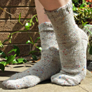 Cozy cream speckled knit socks on feet outdoors, with green plants and brick wall in background. Sunlight creates a warm, inviting atmosphere. Winwick Mum | Basic 6ply Socks | Beginner sock knitting | Free sock knitting patterns