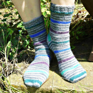 Person wearing striped wool socks with shades of blue, gray, and purple, standing outdoors on a sunlit stone surface surrounded by greenery. Winwick Mum | Basic 8ply DK Socks | Beginner sock knitting | Free sock knitting patterns