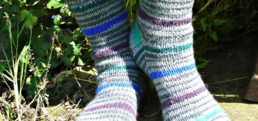 Person wearing striped wool socks with shades of blue, gray, and purple, standing outdoors on a sunlit stone surface surrounded by greenery. Winwick Mum | Basic 8ply DK Socks | Beginner sock knitting | Free sock knitting patterns