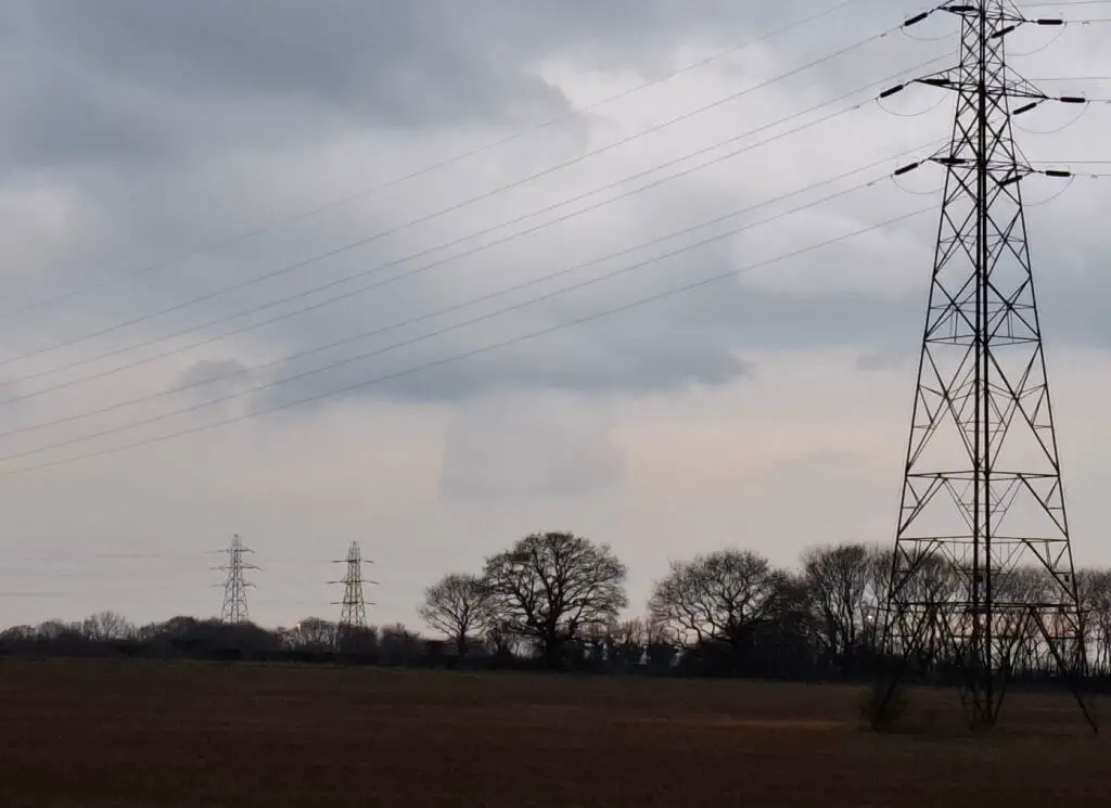 Overcast sky with scattered clouds above a flat field. Three tall electricity pylons dominate the scene, stretching wires across, evoking a calm, industrial landscape.