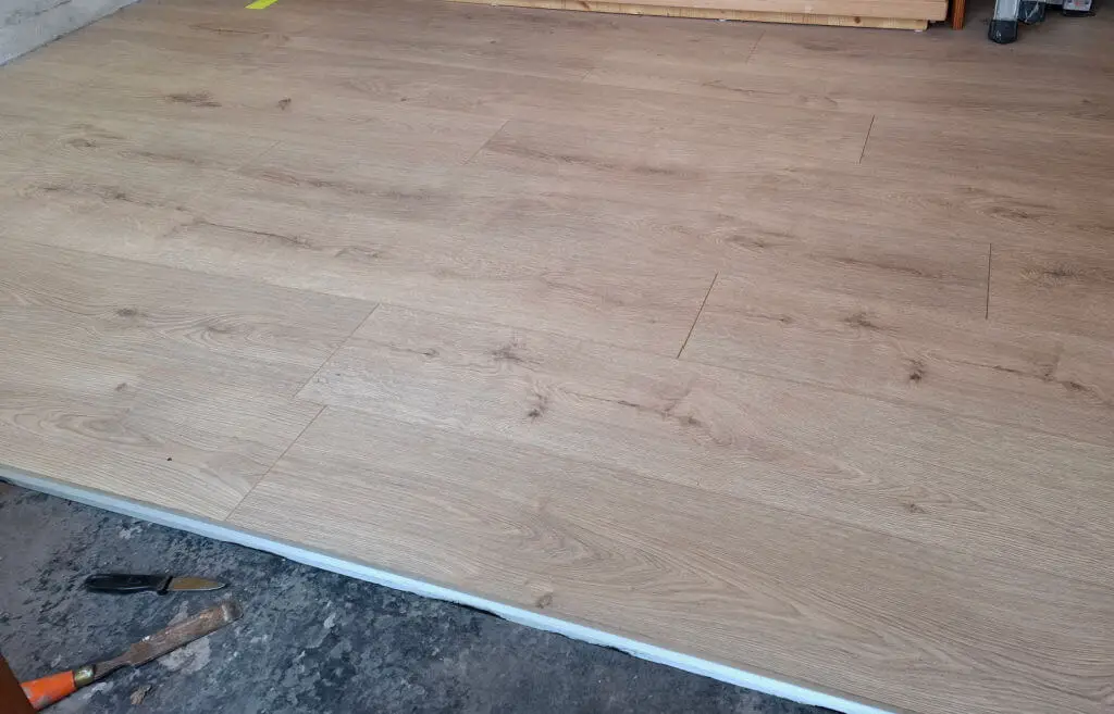 Light oak laminate flooring being installed, partially covering a concrete floor. A chisel and knife are on the concrete, suggesting recent work.