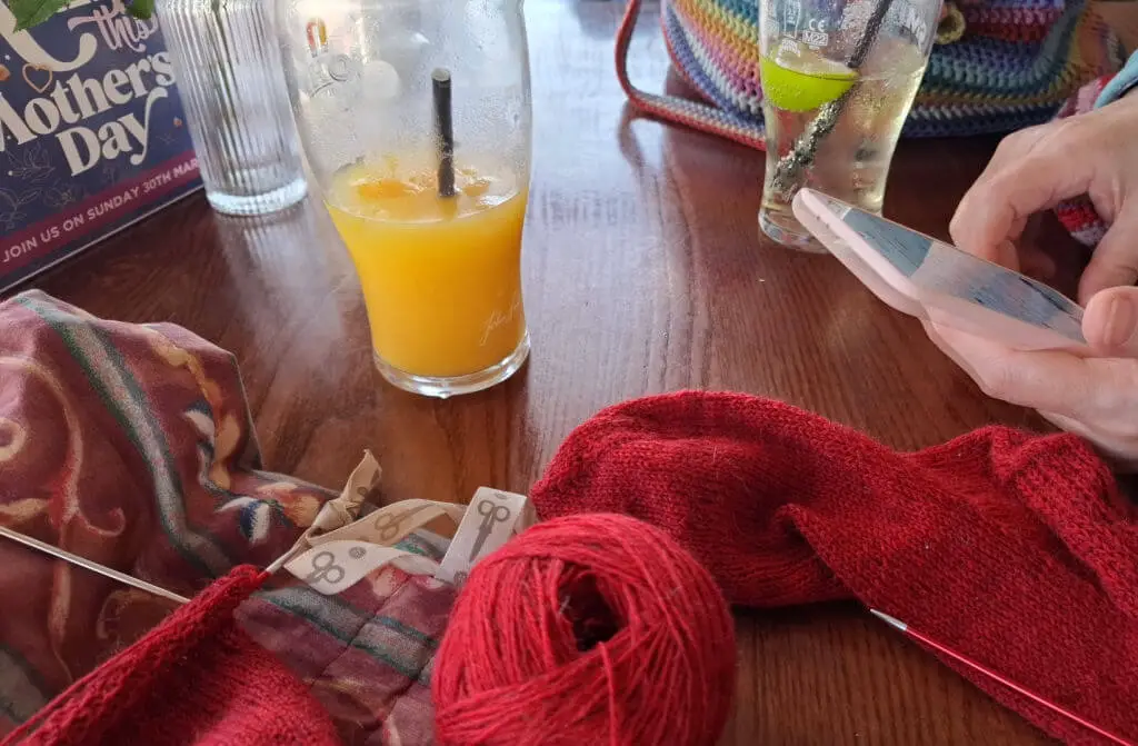 A table with knitting supplies, including red yarn and a scarf, next to a glass of orange juice. A person uses a smartphone, conveying relaxation.